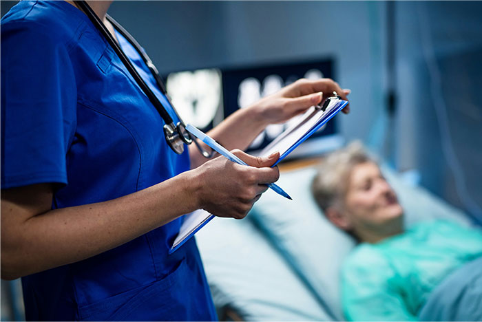 Doctor in blue scrubs holding a clipboard, taking notes while a patient lies in a hospital bed in the background.