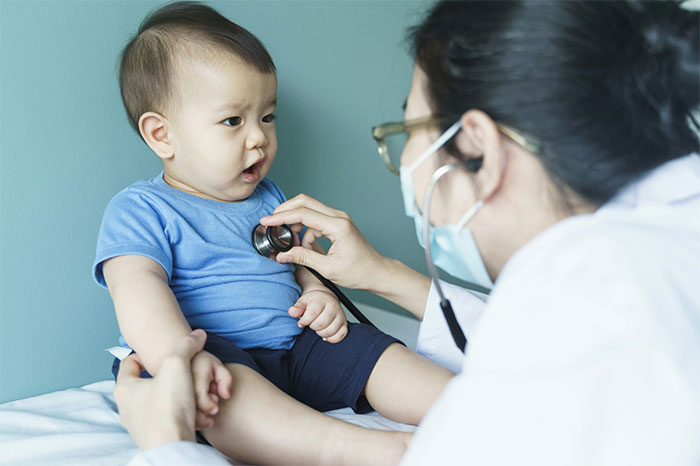 Doctor using stethoscope to examine curious baby boy with medical mask in clinic, highlighting patient and doctor interaction.