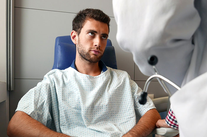 Young male patient in hospital gown looking skeptical while doctor talks, illustrating patients trying to fool doctors.