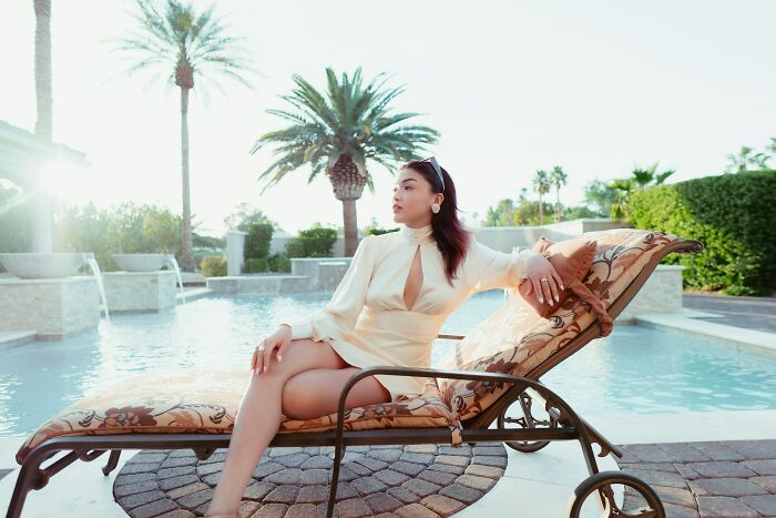 Woman relaxing by pool on lounge chair, reflecting on life and school bullies, surrounded by palm trees and sunlight.