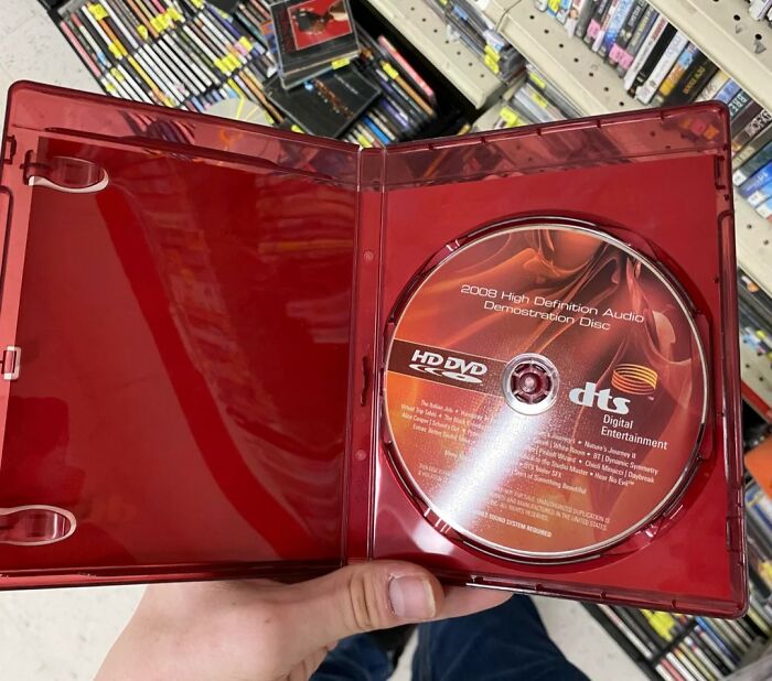 HD DVD disc in red case held in hand in a store aisle filled with DVDs, symbolizing the next big thing that totally flopped.