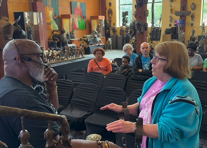 A white woman reacts dramatically while shackled during an African-American museum visit, surrounded by seated visitors.