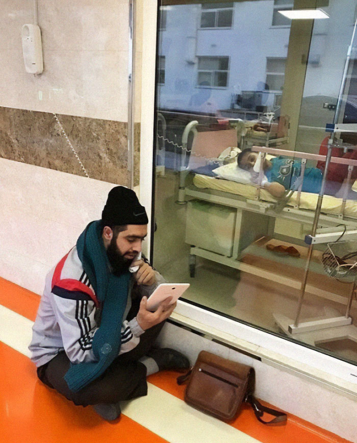 Man sitting on the floor using a phone to video call a patient through a hospital room window, wholesome post for distraction.