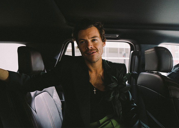 Harry Styles sitting inside a car wearing a black jacket and a cross necklace, casually smiling at the camera.