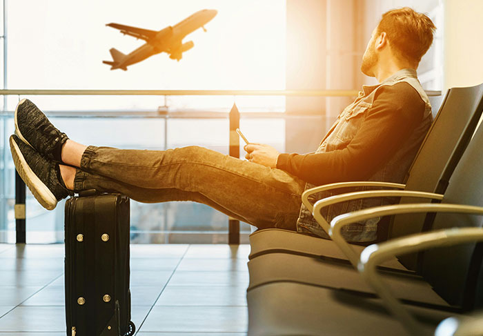 Man sitting in airport waiting area with feet on suitcase watching airplane take off, missing flight home scenario.