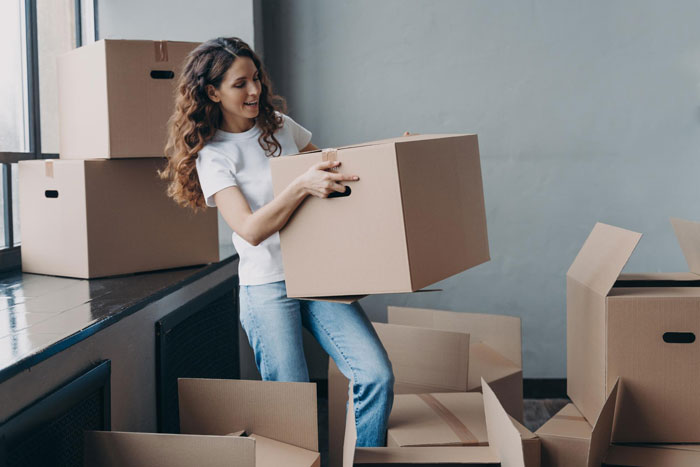 Woman lifting a cardboard box while surrounded by moving boxes, illustrating a wife turned mean once got money scenario.