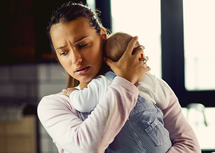 Worried mom holding baby closely inside a home, dealing with parenting and husband nursery ban challenges. Worried mom holding baby closely inside a home, dealing with parenting and husband nursery ban challenges.