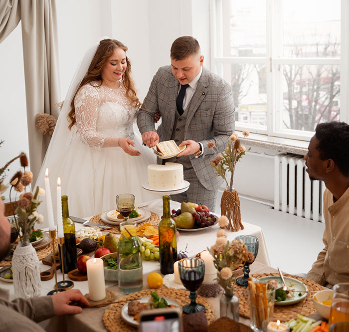 Bride and groom cutting wedding cake while guests sit at table full of food and drinks during celebration