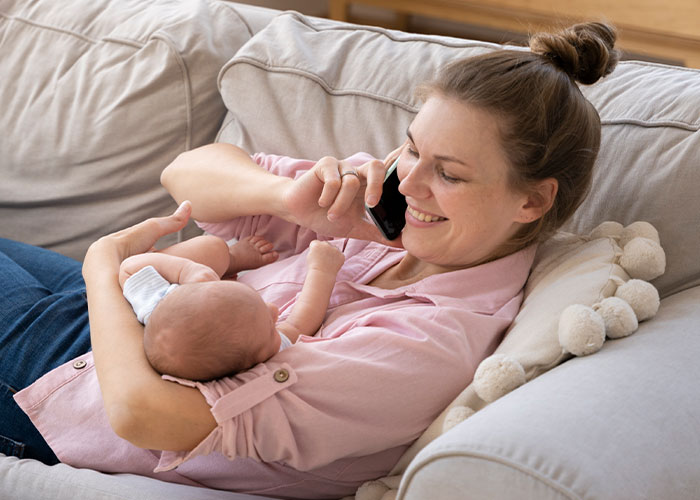 Woman holding baby on couch, smiling while talking on phone, representing affair baby discovery during WIC profile setup.