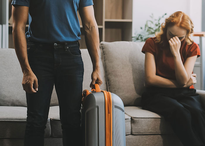 Woman upset on couch after discovering affair baby, man standing nearby holding suitcase in a tense home setting.
