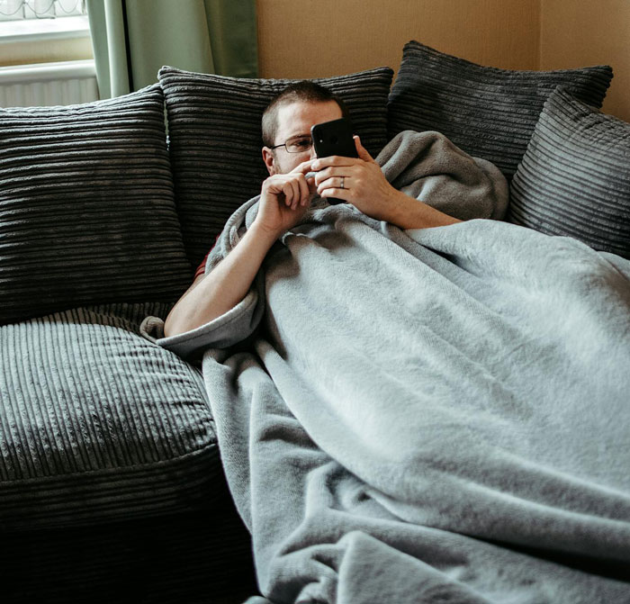 Man wrapped in a blanket on a gray couch, using a smartphone, reflecting on a toxic marriage and family issues. Man wrapped in a blanket on a gray couch, using a smartphone, reflecting on a toxic marriage and family issues.
