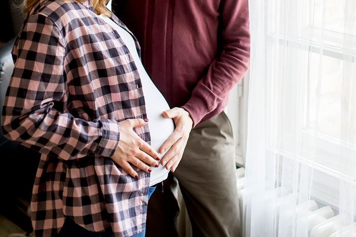 Pregnant woman and partner standing by window, hands on belly, symbolizing desire for motherhood and insemination plans.