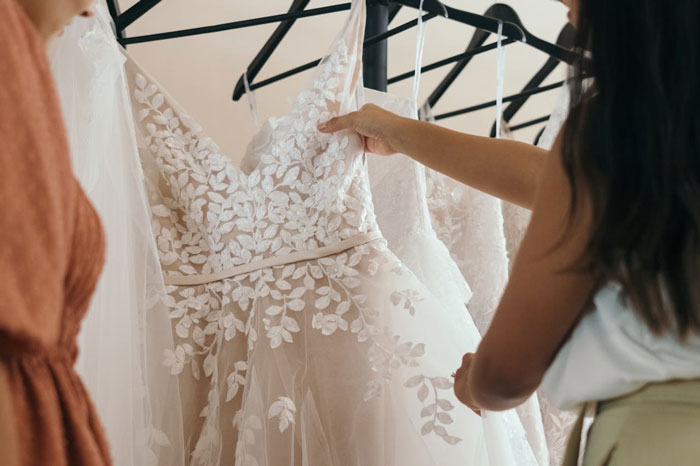 Two women examining white floral lace wedding dresses while dress shopping without the man&rsquo;s fianc&eacute;e present.