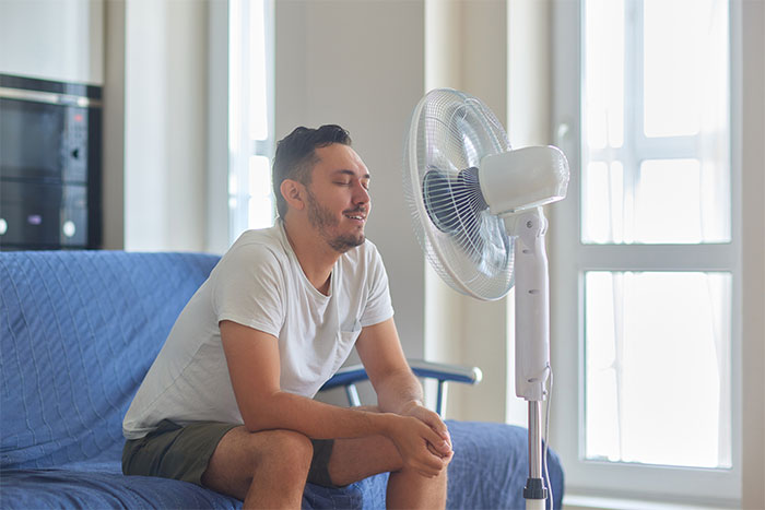 Man sitting on couch cooling off with a fan at home, illustrating living with men experiences shared by women.