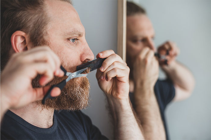 Man grooming his beard with scissors and comb in front of a mirror, highlighting daily living habits of men.