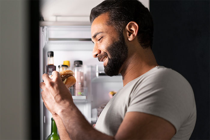 Man smiling while holding a sandwich near an open refrigerator, illustrating living with men insights from women shared.
