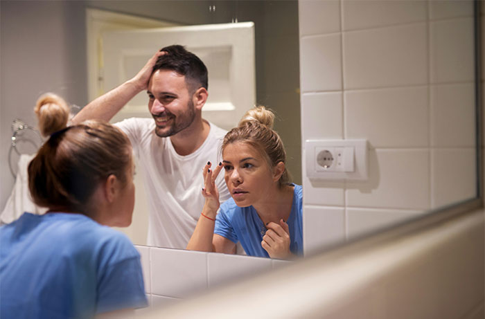 Woman examining her face in the mirror while a man smiles behind her, illustrating women's revelations after living with men.
