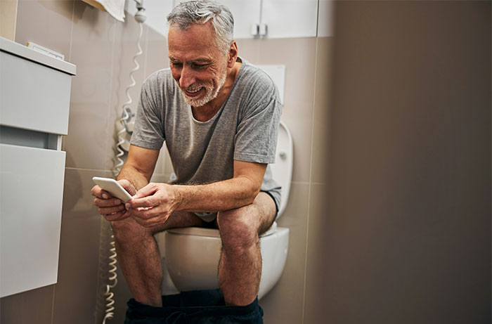 Older man sitting on toilet, smiling and using smartphone, representing daily life moments living with men.