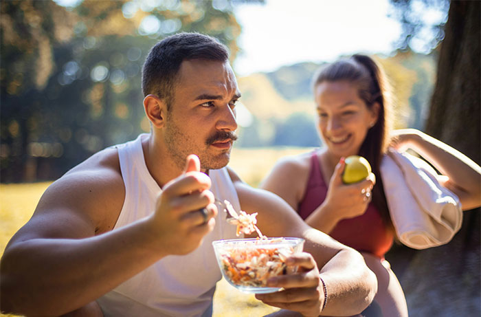Man eating salad outdoors while woman smiles nearby, illustrating insights about living with men shared by women.
