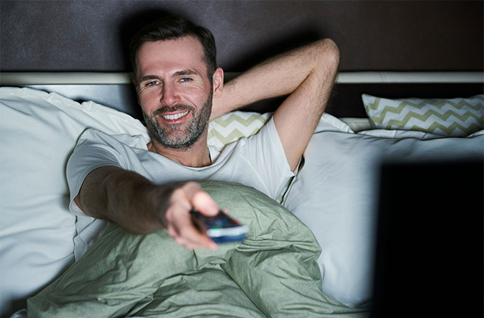 Man relaxing in bed smiling and holding a remote control, illustrating women’s revelations after living with men.
