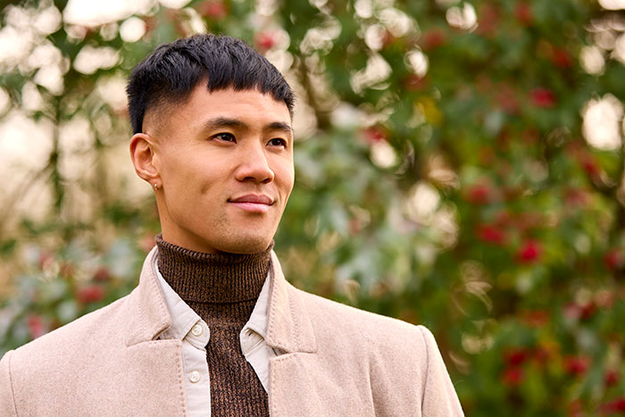 Young man in a beige coat standing outdoors with a confident expression amid blurred greenery and red berries background.
