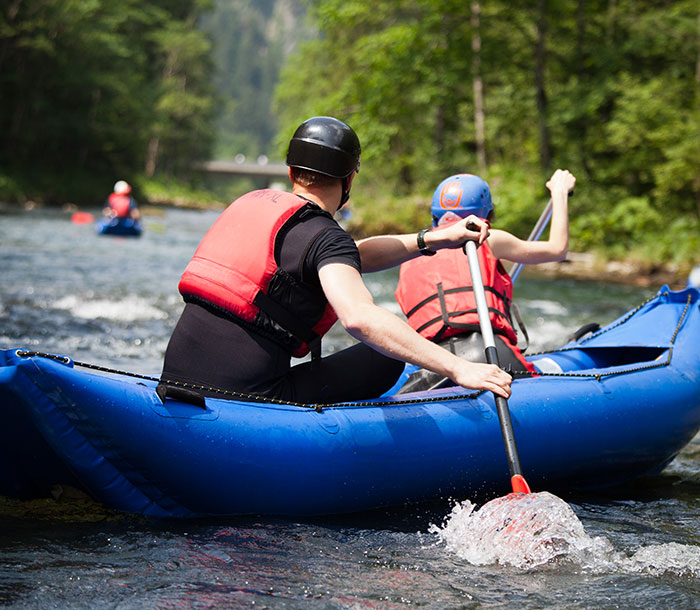 Two people in life jackets and helmets paddling a blue raft on a river surrounded by green forest outdoors.