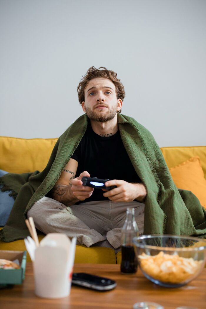 Young man looking grumpy and annoyed while playing video games, wrapped in a green blanket on a yellow sofa.