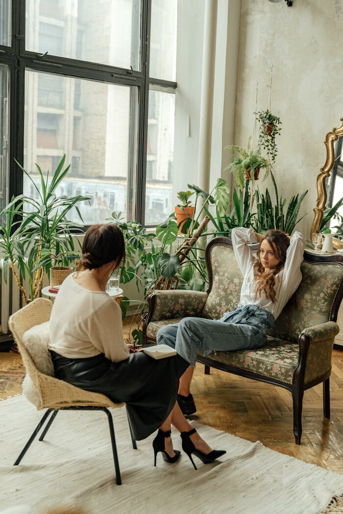 Two women in a living room with plants, one looking grumpy and annoyed while the other listens attentively.
