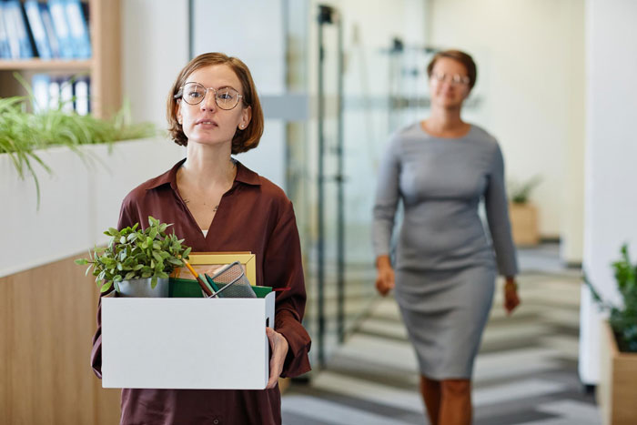 Employee carrying a box of personal items at work, symbolizing being sidelined, with a blurred coworker in the background.