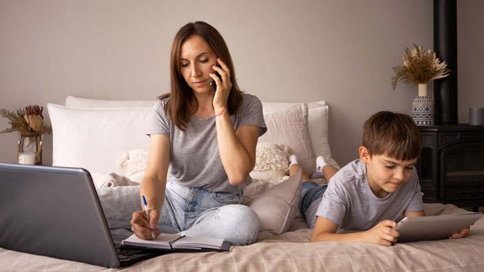 Teen helping with babysitting half siblings while mother talks on phone and works on laptop in cozy bedroom.