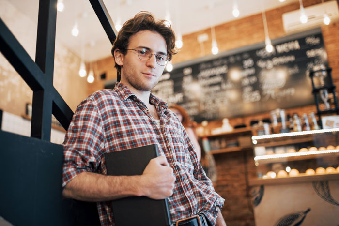 Teen holding a book, leaning against a wall in a caf&eacute;, reflecting on babysitting half siblings and secret plans.