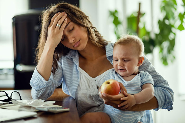 Tired woman holding a baby at a table, appearing stressed while caring for young half siblings and managing babysitting duties.