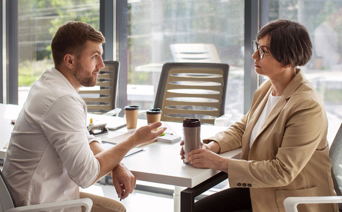 Man and woman having a serious discussion in office while woman holds a travel tumbler during a Tenerife vacation debate.