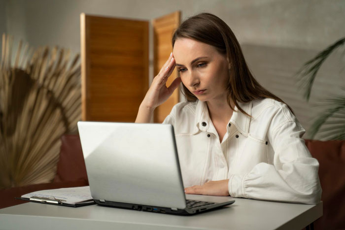 Woman working on laptop at home, appearing stressed as her Tenerife vacay is blocked by boss over focus concerns.