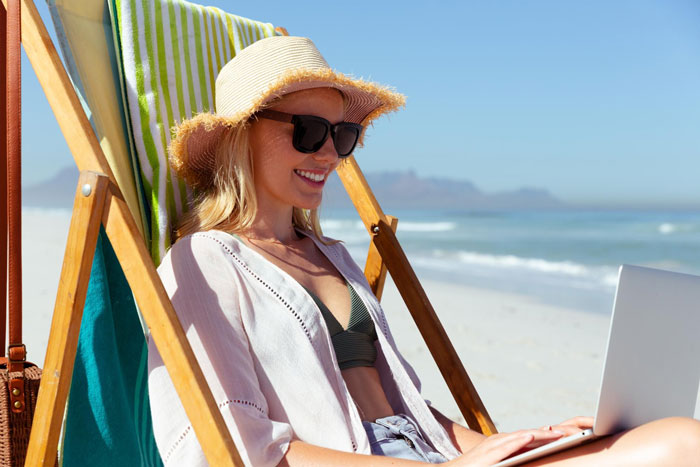 Woman enjoying Tenerife beach while working on laptop, wearing sunhat and sunglasses, blocked from vacation by boss over focus concerns