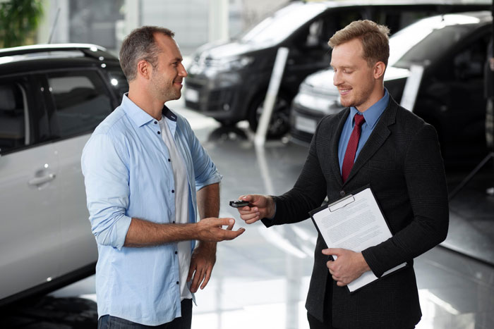 Man in casual wear receiving luxury vehicle keys from salesman in a dealership, highlighting hubby prioritizes luxury vehicle over family plans.