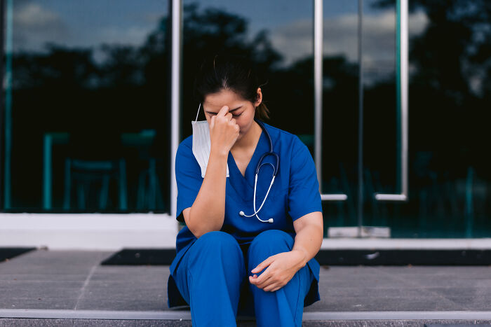 Nurse in blue scrubs sitting outside, holding her head in distress, representing haunting experiences of doctors and nurses.