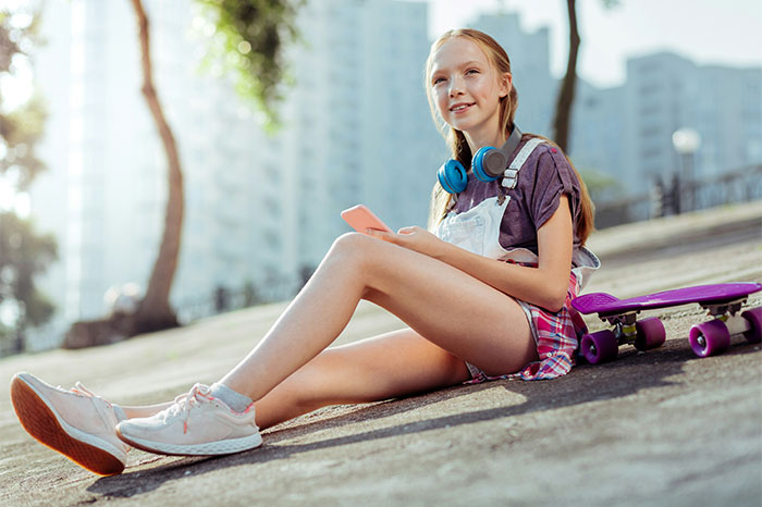 Teen girl sitting outdoors on pavement with skateboard nearby, using phone, representing ways people got banned from a friend’s house.