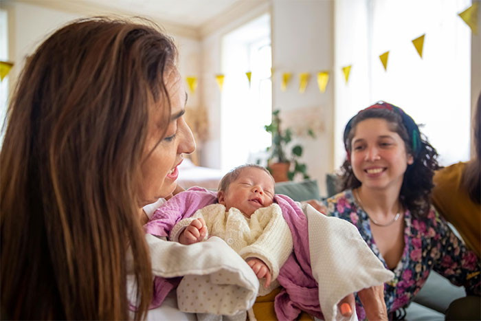 A woman holding a baby while people smile in a cozy living room, illustrating ways people got banned from a friend’s house.