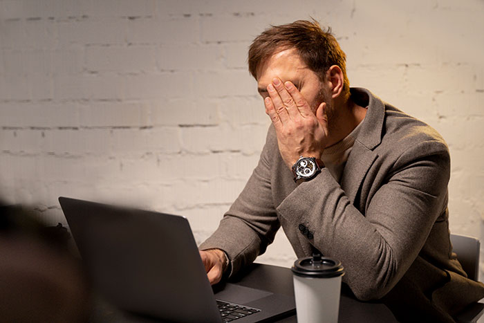 Man in a blazer covering his face in frustration while working on a laptop, illustrating work mess-up stress and recovery.