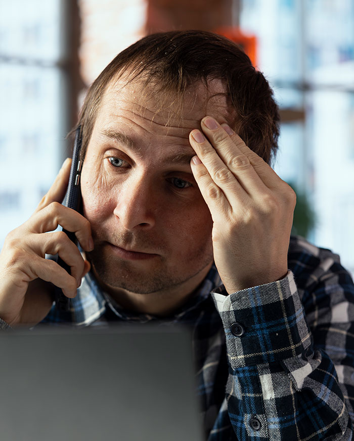 Stressed man on phone, holding forehead, looking at laptop, illustrating people badly messed up at work moments.