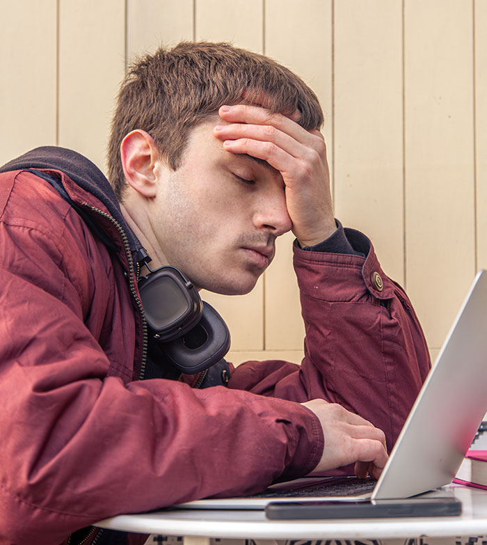 Young man stressed and frustrated at work, wearing headphones around neck, resting head on hand while using laptop.