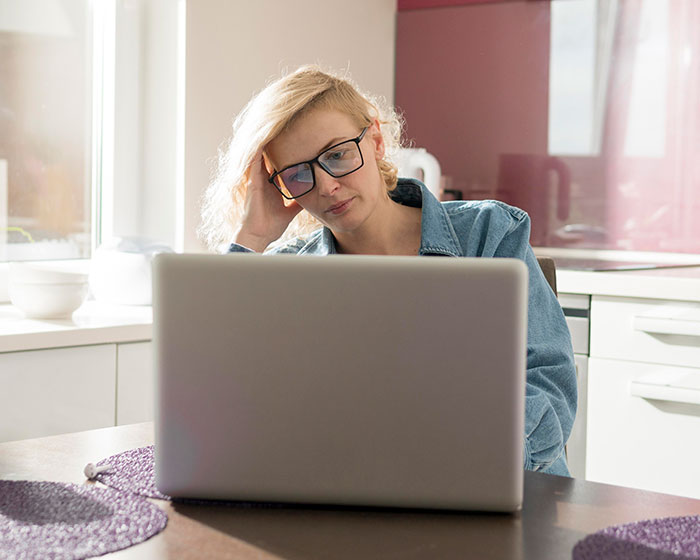 Woman with glasses looking frustrated at laptop in bright kitchen, reflecting moments people messed up at work and lived.