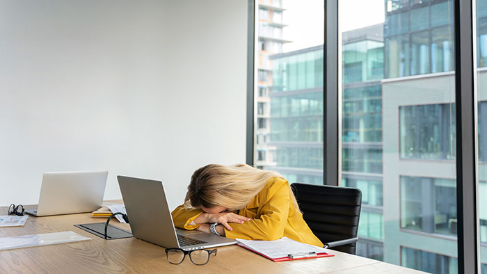 Woman in yellow jacket resting head on arms at desk with laptop, showing exhaustion after work mistake.