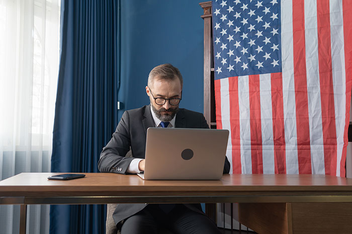 Man in a suit focused on laptop, sitting at desk with American flag, illustrating work mishaps at office environment.
