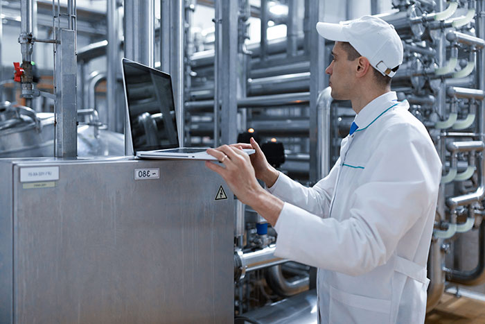 Worker in white coat and cap using laptop in industrial facility representing people badly messed up at work.