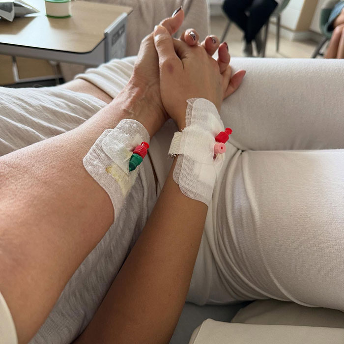 Two hands with IV lines clasped together on a hospital bed, showing support as Bella suffers with darkness and pain.
