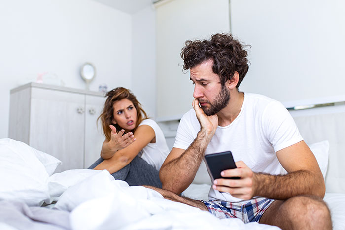 Upset woman confronts man holding phone in bed, illustrating tension with cheater ex in a relationship conflict. Upset woman confronts man holding phone in bed, illustrating tension with cheater ex in a relationship conflict.