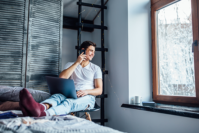 Young man with laptop sitting on bed, smiling while talking on phone during a relaxed hospital visit prank setting.