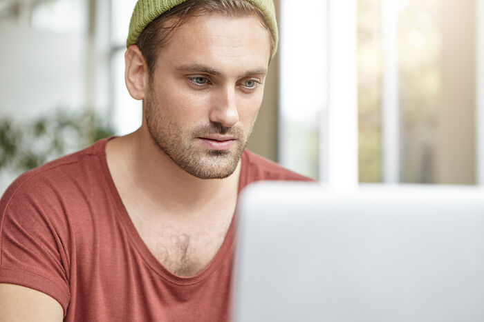 Man in a red shirt and beanie focused on laptop, representing employees sharing insider tricks to simplify jobs.